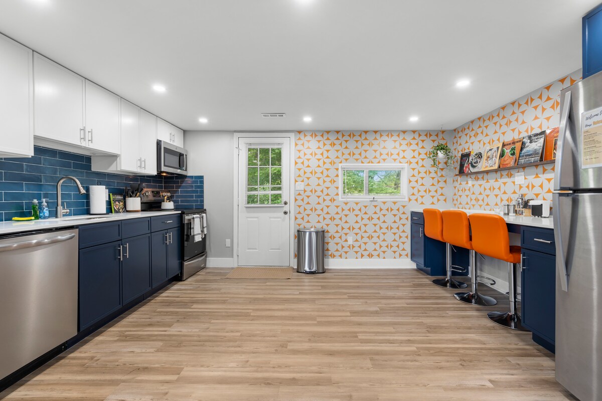 A modern kitchen features sleek navy blue cabinetry paired with stainless steel appliances. An orange accent wall adds a pop of color, while three orange bar stools line the breakfast bar. Natural light filters through a window, illuminating the clean and organized space.