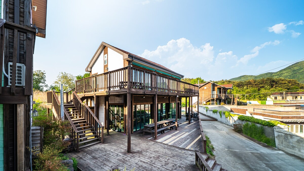 A two-story building is surrounded by wooden decking, featuring stairs leading to an elevated porch. Large windows invite natural light into the structure. Lush greenery and neighboring buildings are visible in the background, with a clear sky enhancing the serene setting.