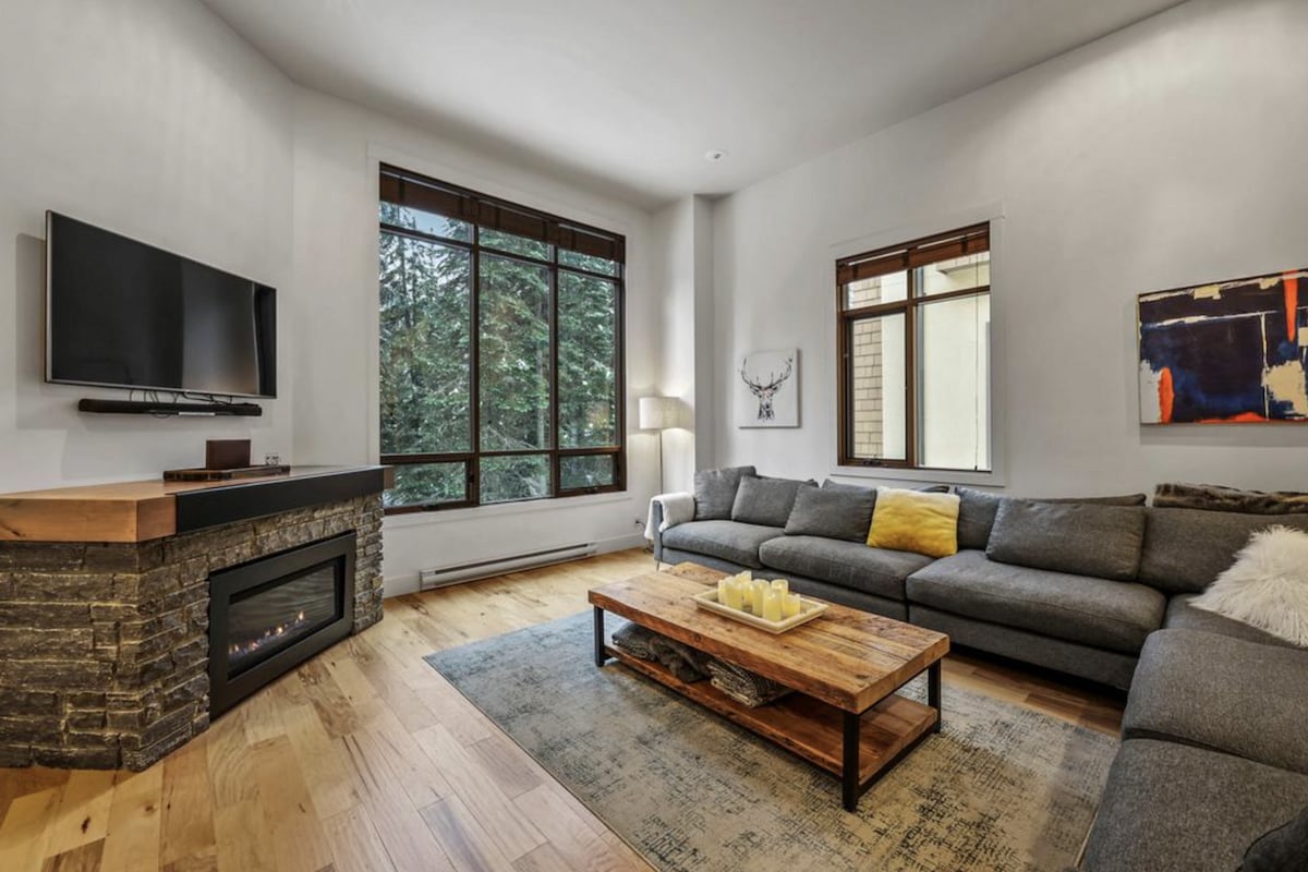 A modern living area is featured, with large windows providing natural light and views of the surrounding greenery. A cozy sectional sofa is positioned around a wooden coffee table, complemented by a stone fireplace. Decorative art pieces add character to the space.