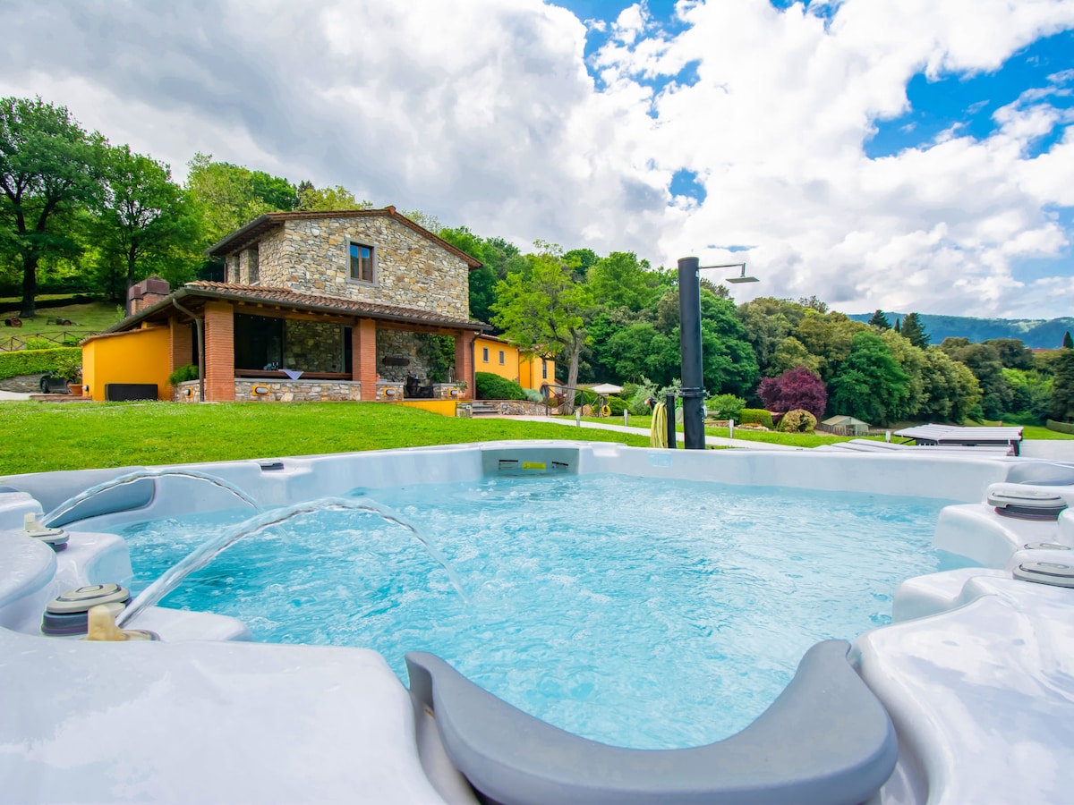 A hot tub is positioned in the foreground, featuring jets that create gentle bubbles. In the background, a three-story farmhouse is visible, surrounded by lush greenery and trees. The sky is partly cloudy, adding depth to the outdoor setting.
