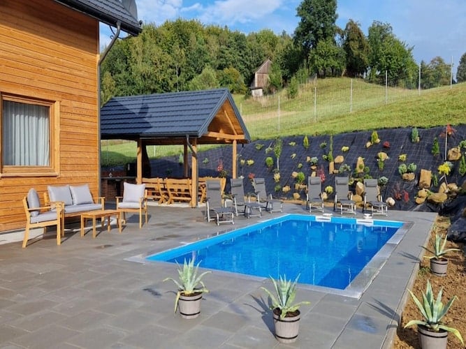 An outdoor area features a private swimming pool surrounded by sun loungers and patio furniture. A barbecue gazebo is visible in the back, while landscaped garden elements, including potted plants, adorn the surrounding space. The setting is framed by a gentle hillside with greenery.