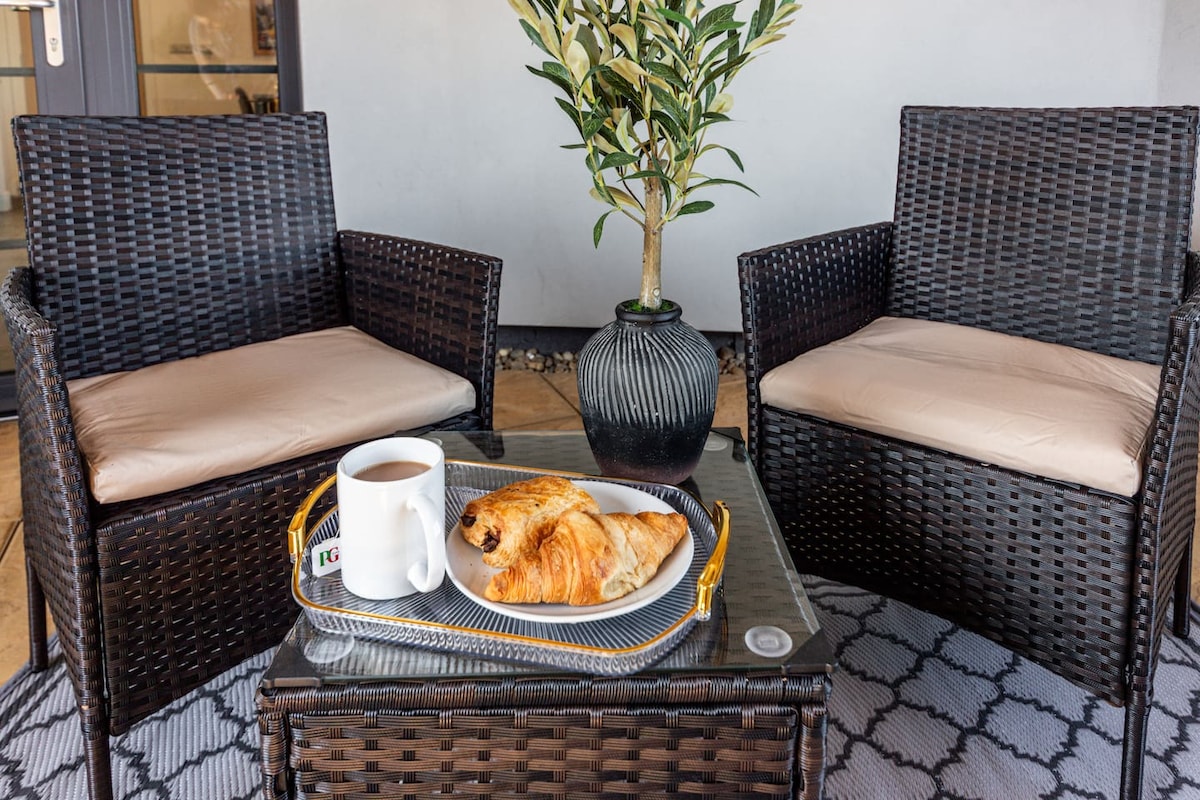 An outdoor seating area is displayed, featuring two woven chairs with soft cushions. A small table holds a tray with a cup of tea and a pastry. A potted plant adds a touch of greenery, enhancing the welcoming atmosphere.