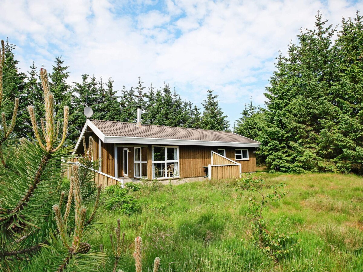 A holiday cottage is nestled in a natural plot surrounded by tall trees. The wooden exterior features large windows that allow for natural light. A small staircase leads to the entrance, while lush green grass and scattered foliage enhance the serene setting.