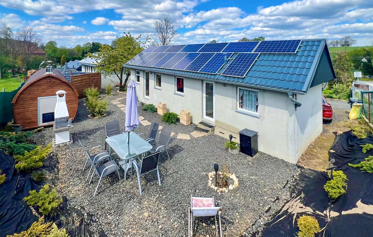 An outdoor area showcases a holiday home surrounded by landscaped grounds. Solar panels are installed on the roof. A glass dining table with chairs is positioned on gravel, near a sun umbrella. A wooden structure and barbecue grill are also visible in the garden.