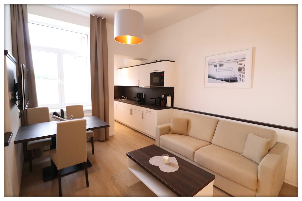 A welcoming living area features a soft sofa and a coffee table at its center. A kitchenette with modern appliances is visible against one wall. A dining table for four is positioned near large windows, with natural light illuminating the space and light curtains enhancing the ambiance.