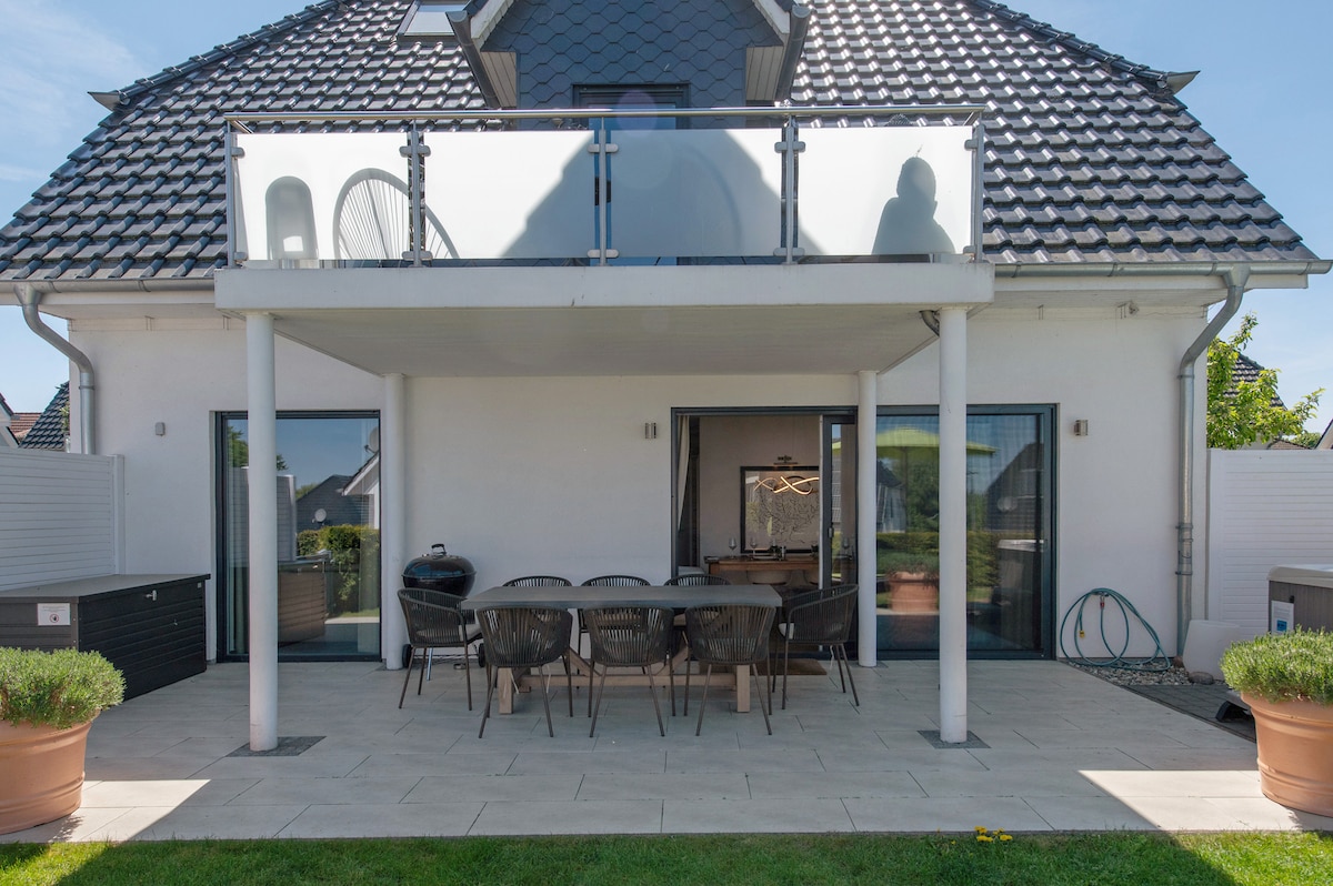 A covered terrace is shown, featuring a large dining table surrounded by several chairs. Glass railings provide a view of the surrounding lawn and garden. A barbecue and a seating area can be seen, with the house’s façade and windows in the background.