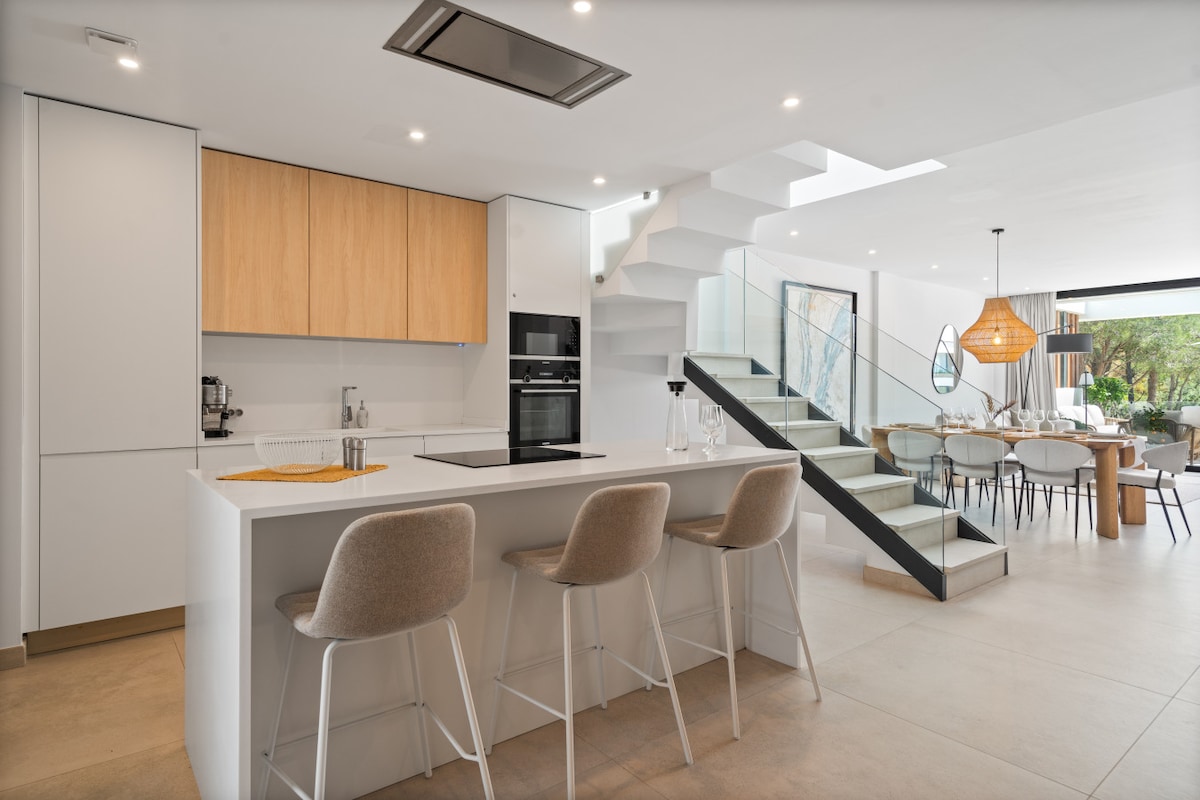 An open space kitchen features a central white island with seating for three. Light wooden cabinets complement modern appliances, while sleek stairs lead to the upper level. A dining area with a large table is visible, surrounded by expansive glass doors showcasing greenery outside.