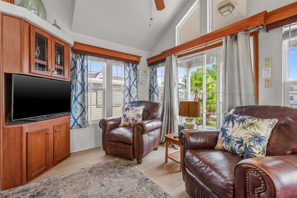 A spacious living area features large windows allowing natural light, with patterned curtains framing the views. Comfortable leather armchairs are positioned near a wooden entertainment unit, which holds a television. A subtle area rug lies beneath, adding a touch of warmth to the space.