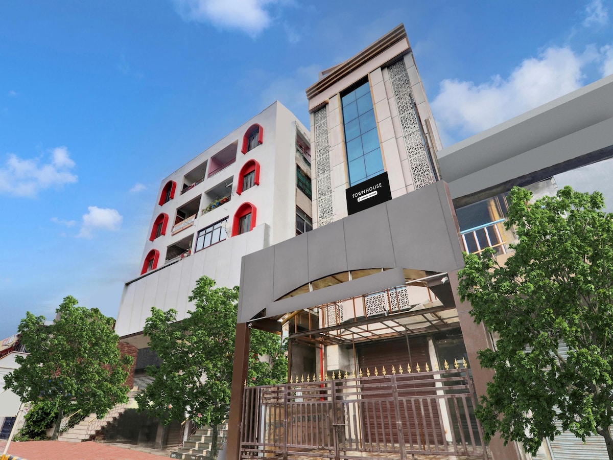 A modern multi-story building is showcased, featuring a prominent entrance with a sleek awning. Brightly colored arch windows are visible, reflecting contemporary design elements. Lush greenery flanks the structure, enhancing the inviting exterior, while a clear sky adds to the overall sense of space.