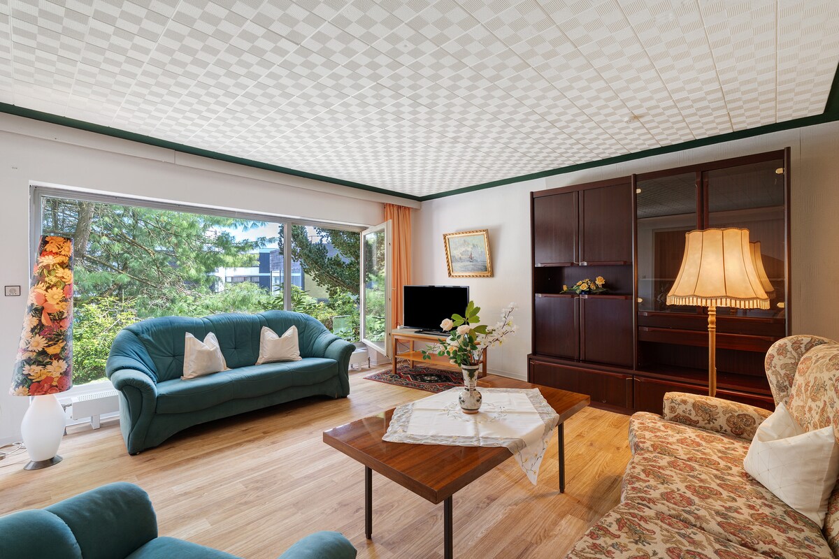 The living room features a spacious seating area with a green couch and a floral patterned sofa. A coffee table is centered between the seating. Large windows provide a view of the surrounding greenery, with a television and wooden cabinet positioned along the wall.