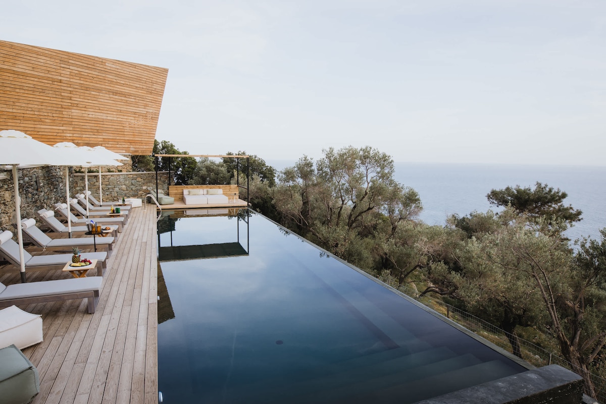 An infinity pool reflects the serene landscape, providing expansive views of the sea. Sun loungers are positioned nearby, accompanied by small tables holding drinks. A wooden deck enhances the outdoor space, with gentle greenery framing the tranquil scene.