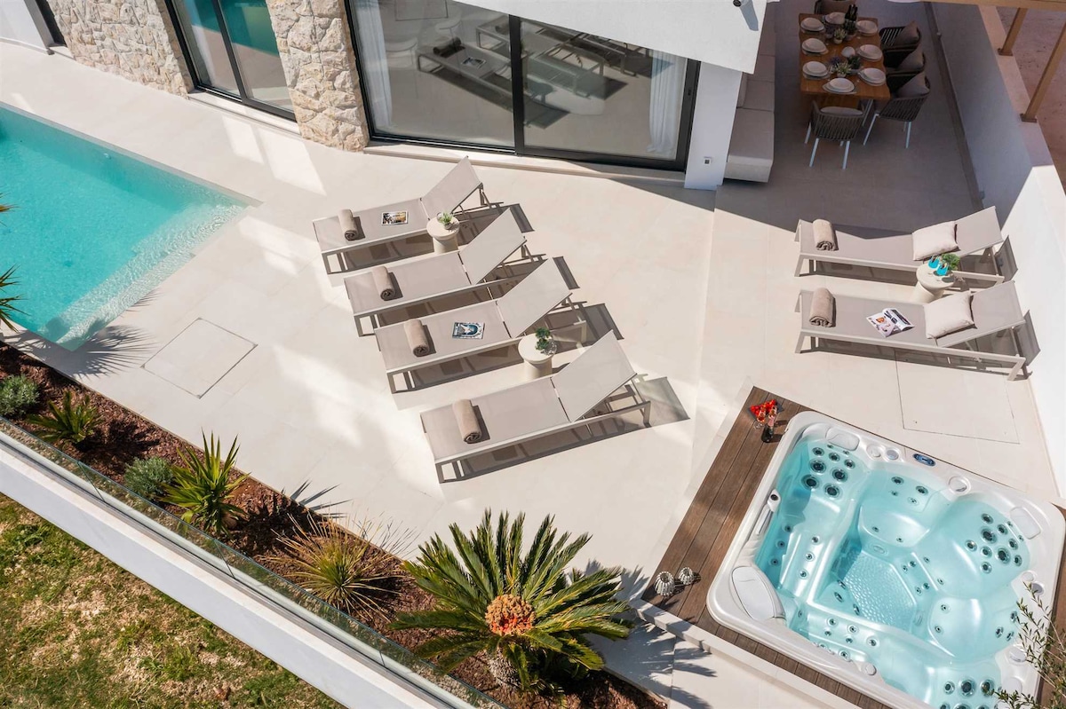 An aerial view showcases the outdoor relaxation area featuring a heated pool and a hot tub. Sun loungers are arranged neatly around the pool, accompanied by small side tables. Lush landscaping adds a natural touch to the elegant outdoor space.