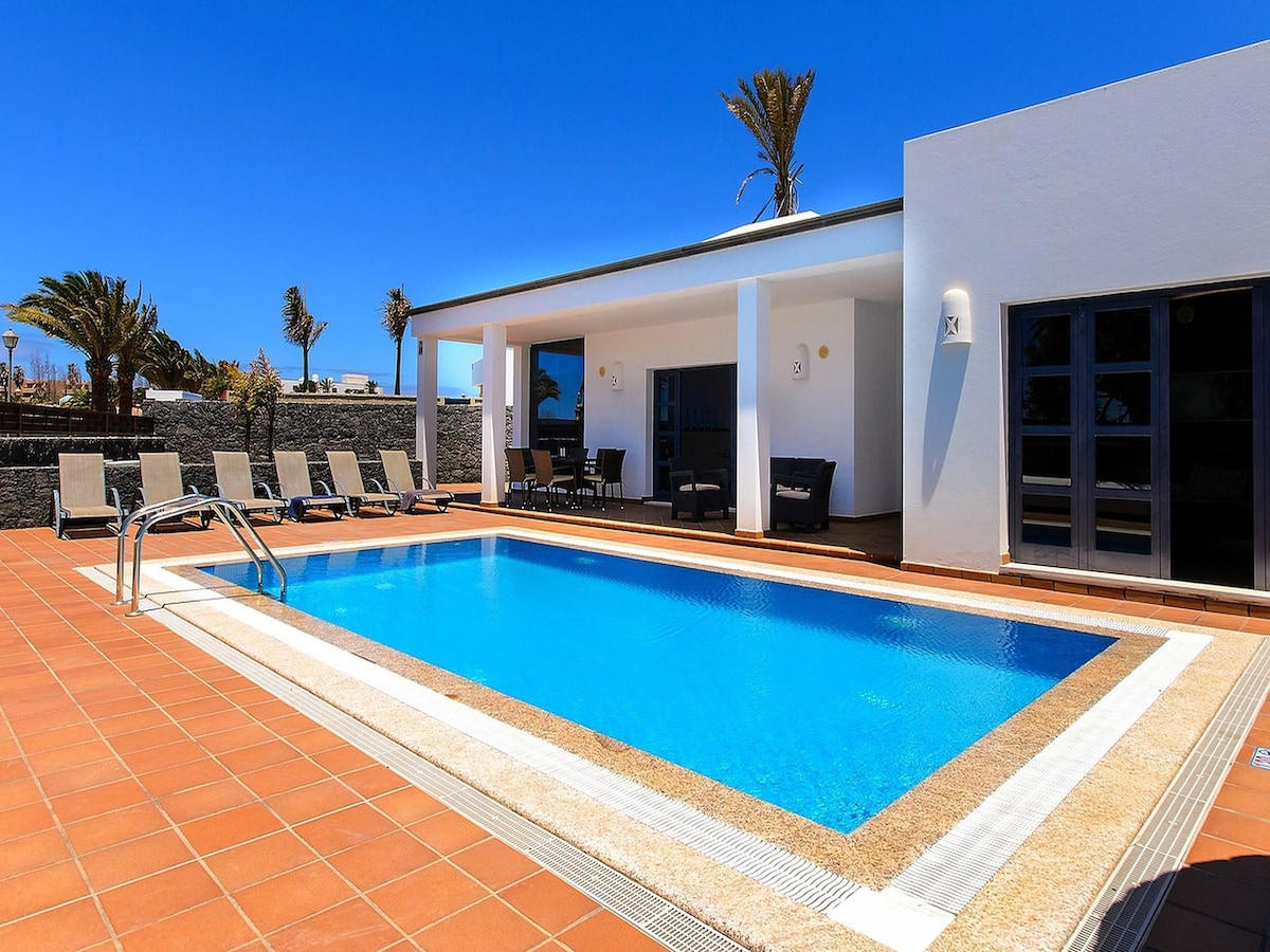An outdoor area features a private swimming pool surrounded by a tiled patio. Several lounge chairs are positioned along the pool, under clear blue skies and palm trees, creating an inviting space for relaxation and sunbathing.
