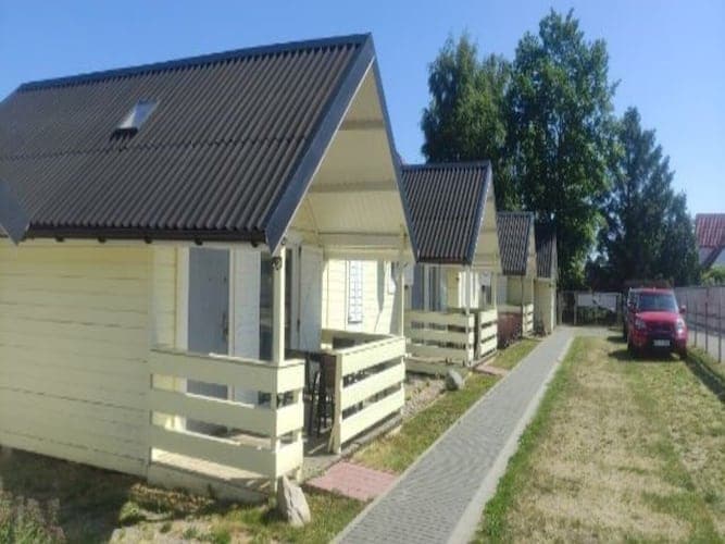 A row of charming holiday cottages is set amidst a grassy area, featuring distinctive sloped roofs. Each cottage has a covered terrace and light-coloured siding, while a neatly paved walkway connects the properties. A parked car is visible to the right, providing convenient access.