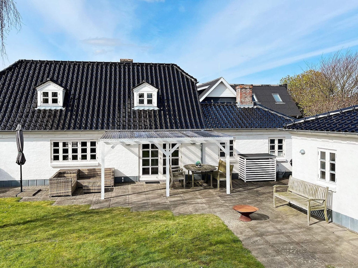 A terrace area is visible, featuring a dining table and chairs beneath a roofed structure. A grassy lawn surrounds the patio, with a fire pit nearby. The white exterior of the holiday home is contrasted by a dark roof, while multiple windows allow natural light into the interior.