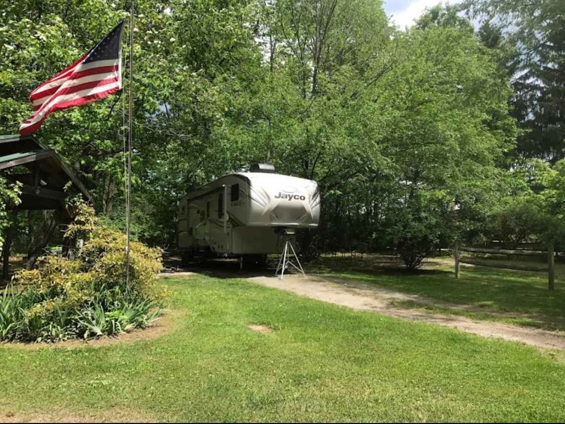 A trailer is positioned on a grassy area, surrounded by lush green trees. A flagpole with an American flag stands nearby, complementing the tranquil setting. The path leading to the trailer is lined with well-maintained foliage.