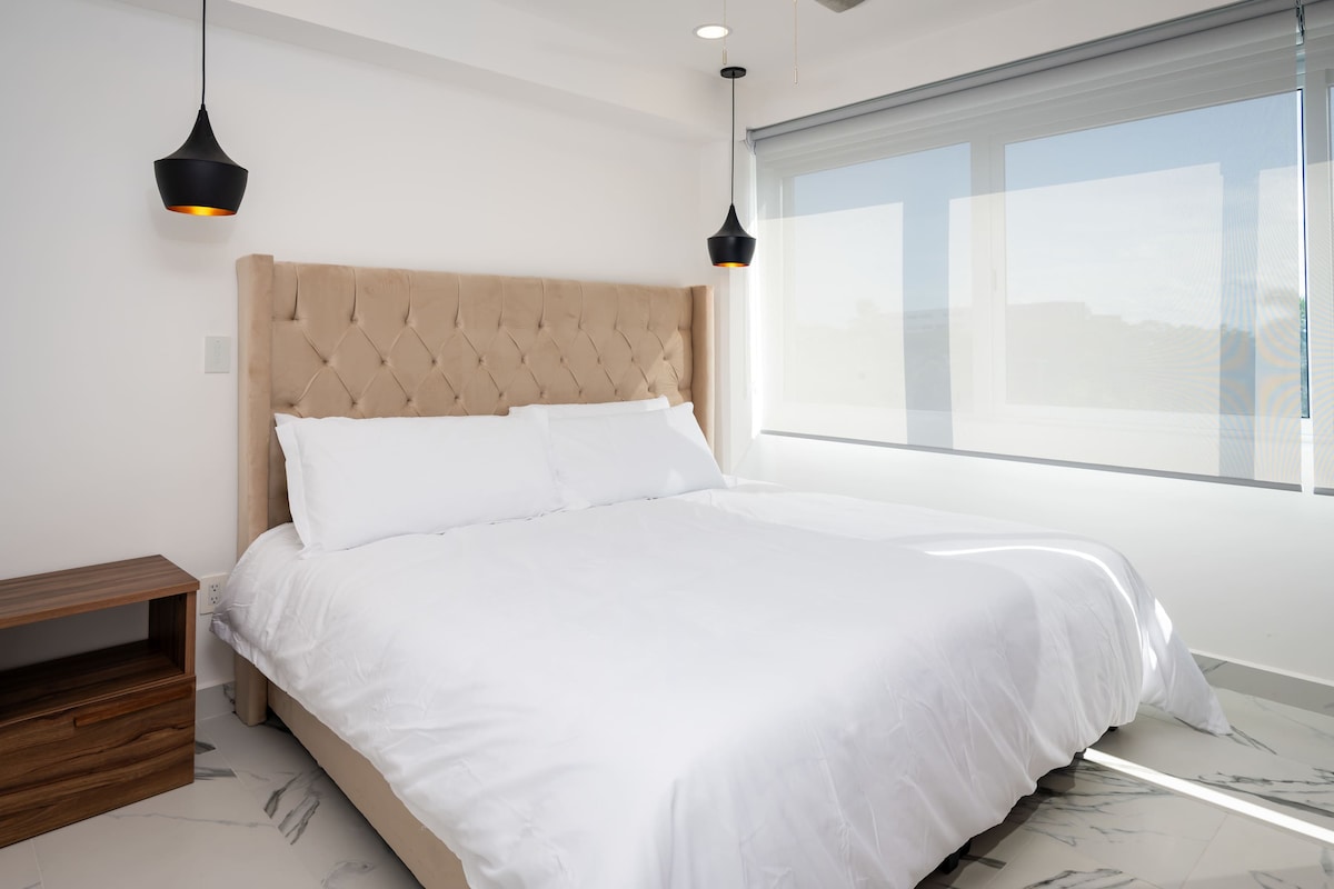 A spacious bedroom is highlighted by a tufted headboard and plush bedding. Two pendant lights hang above, providing soft illumination. Large windows allow natural light to fill the room, while a bedside table offers additional storage.