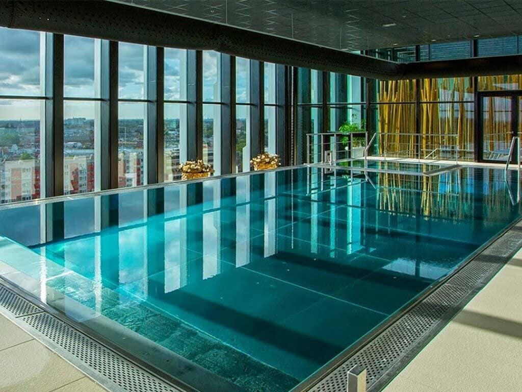 An indoor pool is showcased with clear blue water reflecting the surrounding floor-to-ceiling windows. The pool's edge features stainless steel detailing, and minimalistic lounge areas are visible nearby, creating a bright and open space.