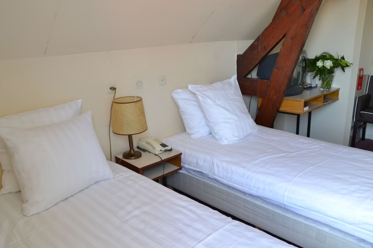 The room features two spacious beds dressed in white linens, positioned against a slanted ceiling with exposed wooden beams. A bedside lamp and a telephone are situated on a wooden nightstand. A small desk is placed nearby, accompanied by a chair.