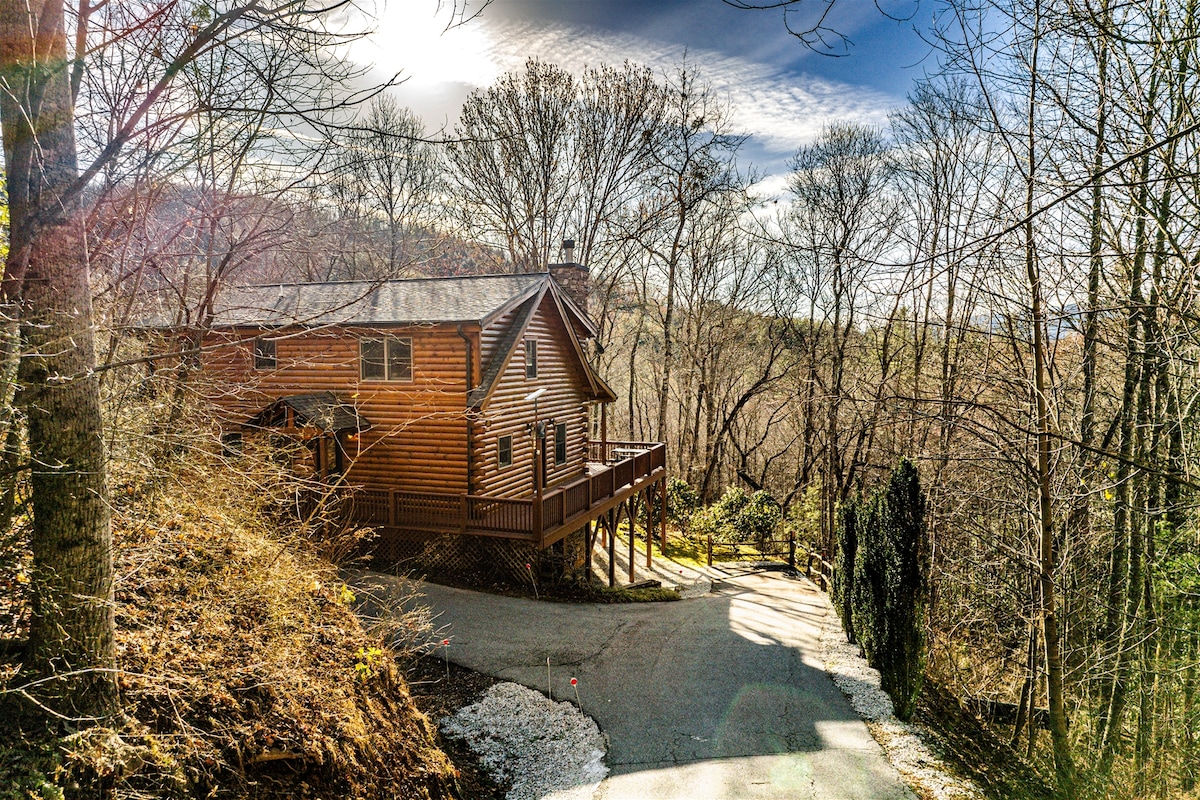A three-story cabin constructed of natural wood is showcased against a backdrop of trees. The expansive deck is visible, providing a welcoming entrance. A gravel driveway winds up to the cabin, bordered by a wooded landscape and a clear blue sky.