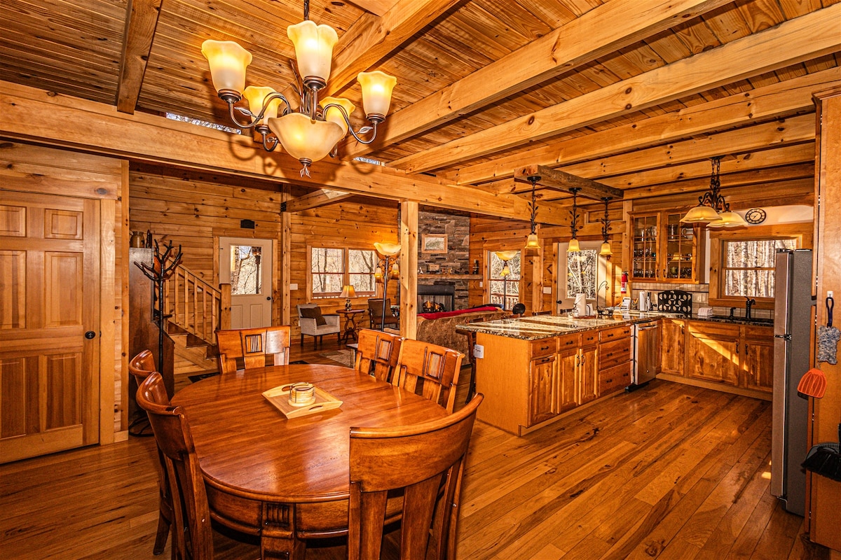 An open kitchen and dining area showcase wooden beams and rustic furnishings. A large table accommodates multiple guests, while the kitchen features ample cabinetry and modern appliances. Natural light filters in from windows, highlighting the warm wood tones throughout the space.