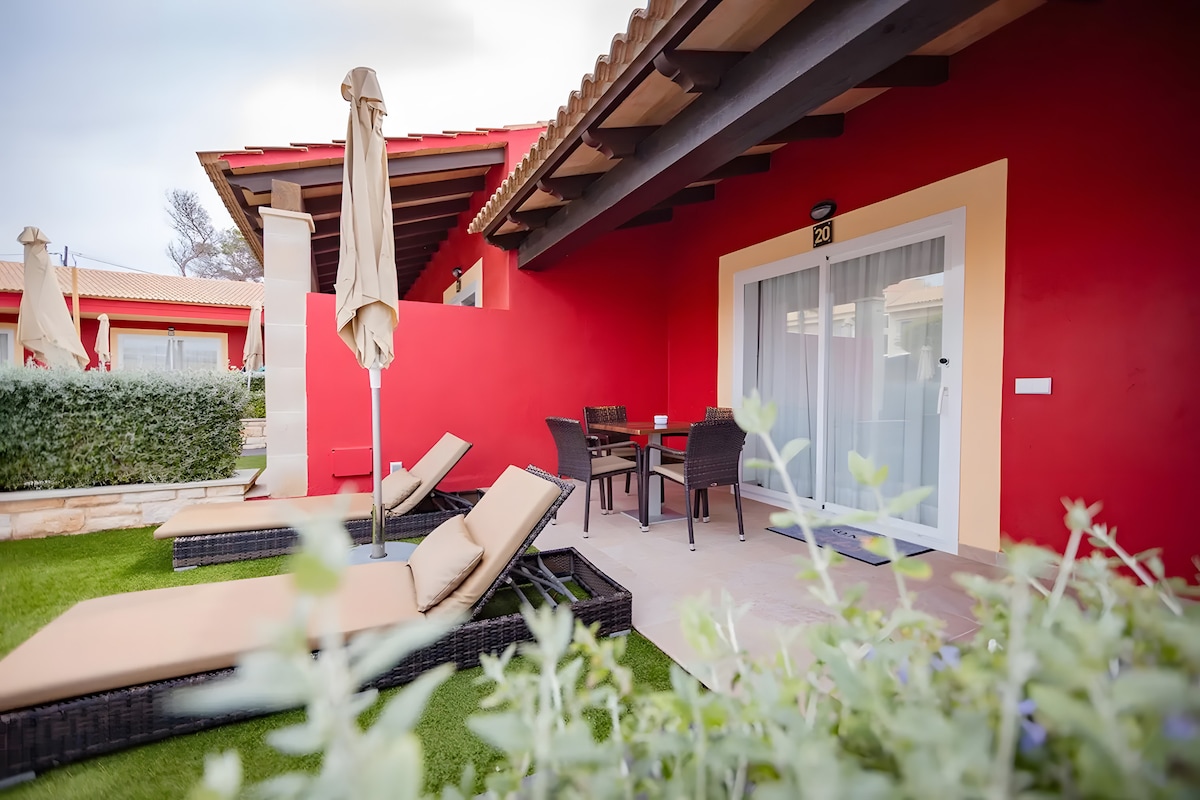 A private covered terrace features outdoor seating with a dining table and chairs. Two lounge chairs are positioned on a grassy area, accompanied by a vibrant red wall backdrop. Umbrellas stand nearby, providing shade and enhancing the inviting space.