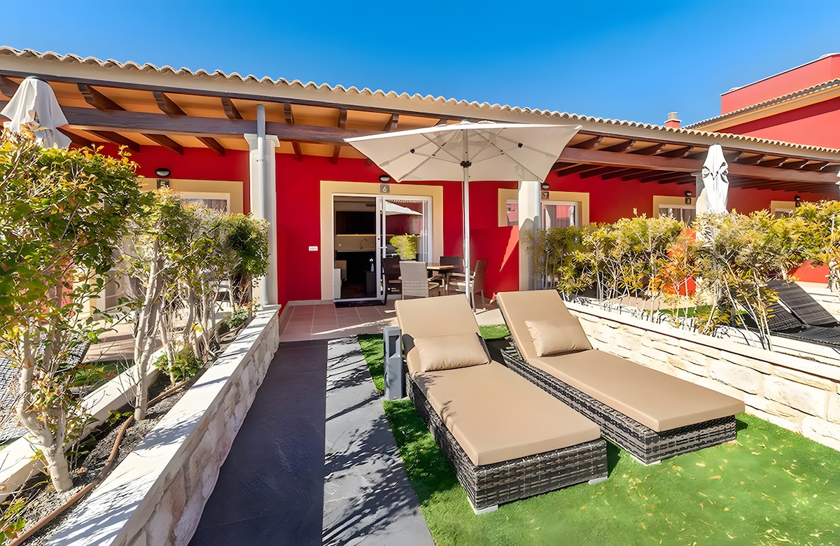 An outdoor terrace features two lounge chairs beside a small table. The area is lined with greenery and has an umbrella for shade. The red exterior walls of the apartment provide a vibrant backdrop, enhancing the inviting outdoor space.