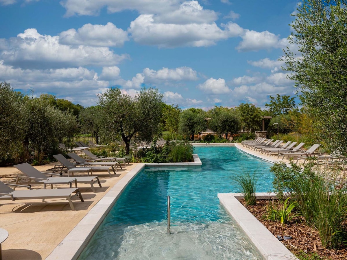 A spacious swimming pool with clear blue water is featured, surrounded by olive trees and landscaped greenery. Lounge chairs are neatly arranged, providing space for relaxation. The atmosphere is bright with a partly cloudy sky adding to the tranquil setting.