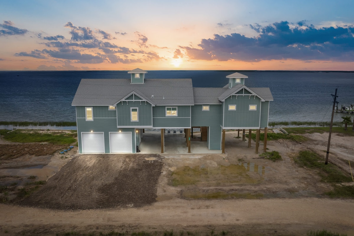 The home is showcased against a sunset backdrop, highlighting its modern design and elevated structure. Large garages can be seen on the ground level, with expansive windows allowing for natural light. The wide expanse of water is visible in the distance, creating a serene coastal setting.