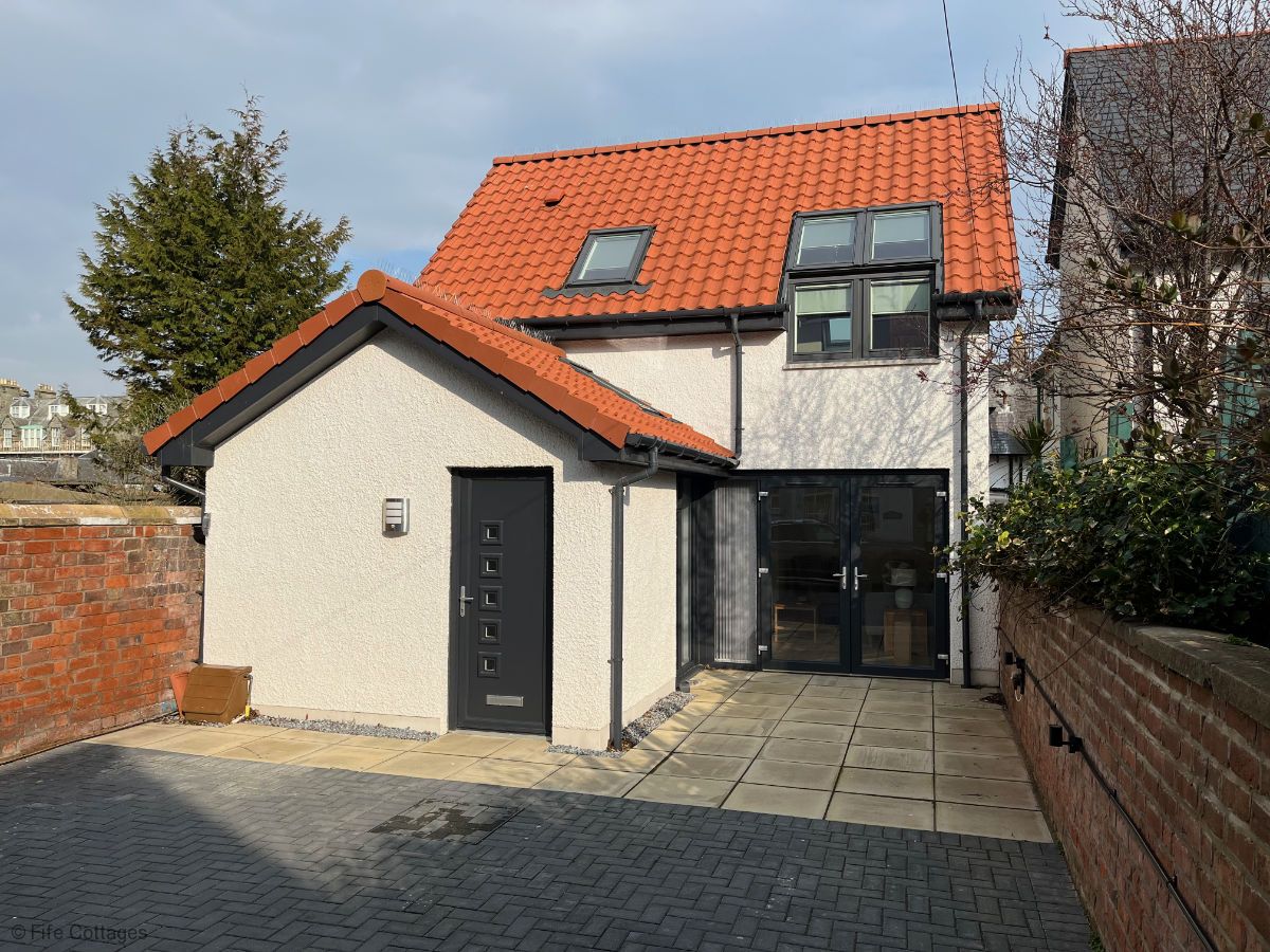 The two-story cottage features a modern design with a red tiled roof. A paved parking area is located in front of the house, providing space for one vehicle. Large windows allow natural light to enter, enhancing the charming exterior.