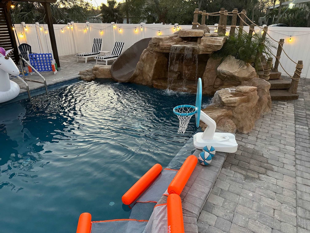 A private pool area features a waterfall and rock formation, accompanied by a basketball hoop. Floating pool loungers and colorful pool toys add to the recreational setting. Reclining chairs and string lights create a relaxed atmosphere around the pool.
