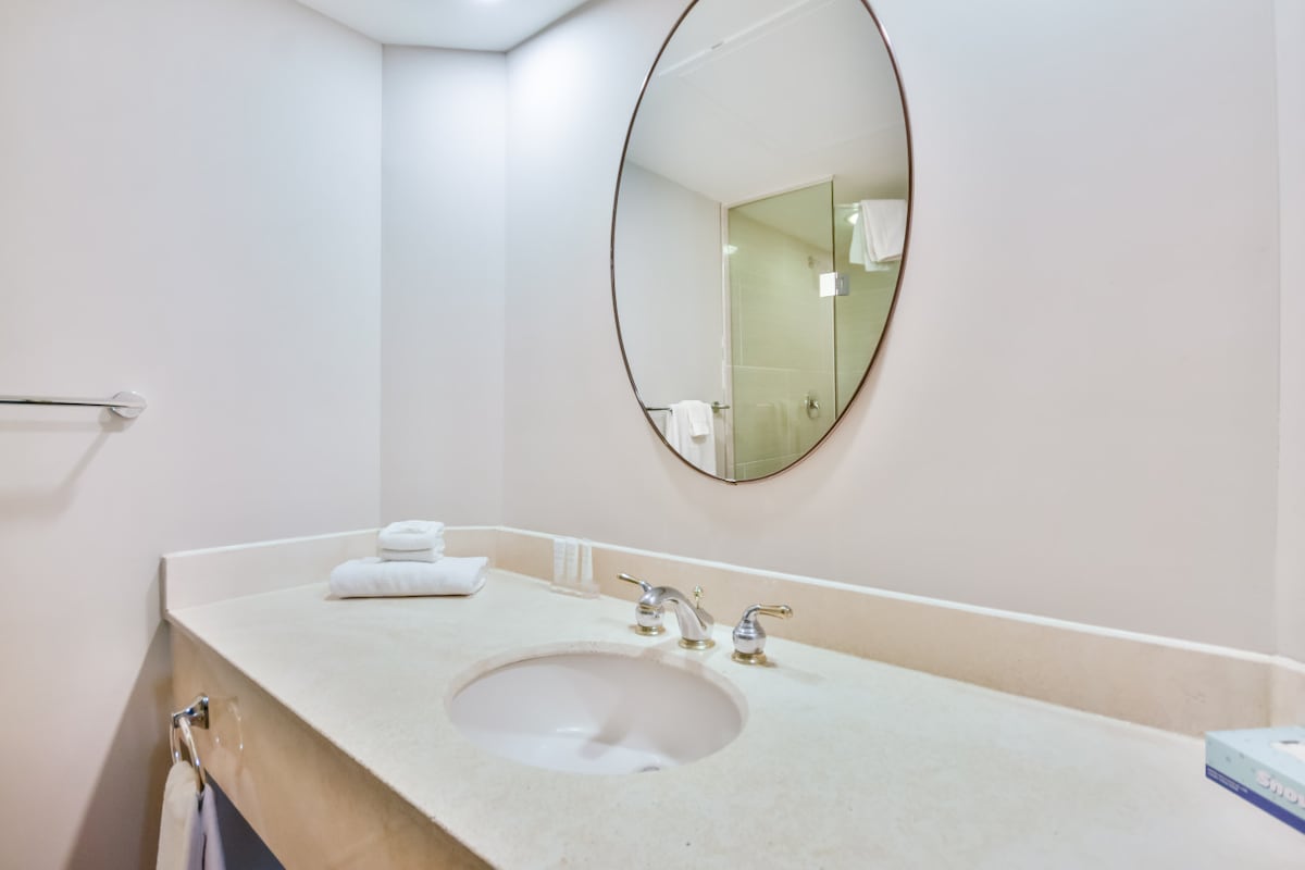 A clean and modern bathroom features a round mirror above a spacious countertop. Neatly stacked towels are arranged beside a single sink, with toiletries placed in a row. A glass-enclosed shower is visible in the background, showcasing a minimalist design.