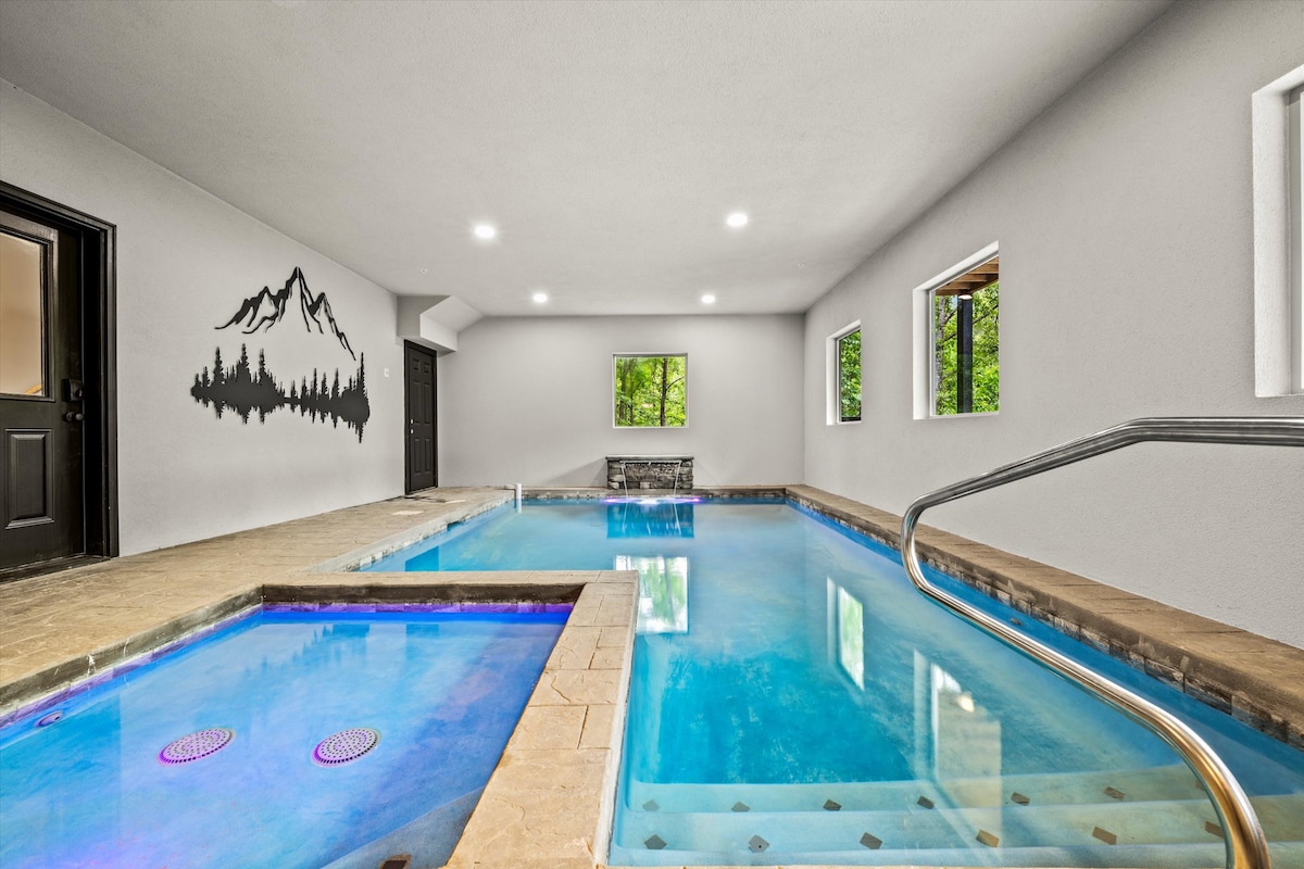 An indoor heated pool area features a large, clear pool accompanied by a separate kiddie pool. Natural light enters through multiple windows, with a mountain silhouette mural adorning the wall. A grab rail is positioned near the pool for safety.