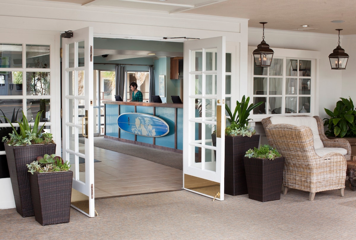 Double doors open to a welcoming lobby area, featuring a reception desk in the background. Planters with greenery line the entrance, while comfortable seating is arranged nearby. Natural light fills the space, enhancing the inviting atmosphere.
