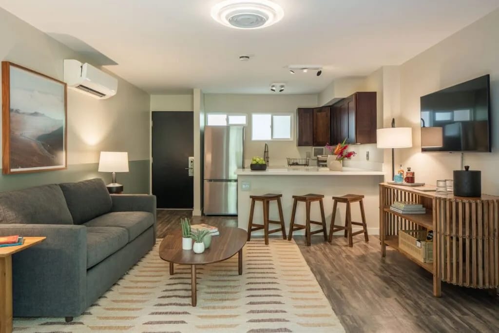 The living area features a dark gray sofa and a round coffee table atop a patterned rug. Two wooden stools are positioned at a kitchen island equipped with modern appliances. A mounted flat-screen TV is visible on the wall, accompanied by natural light from the nearby windows.