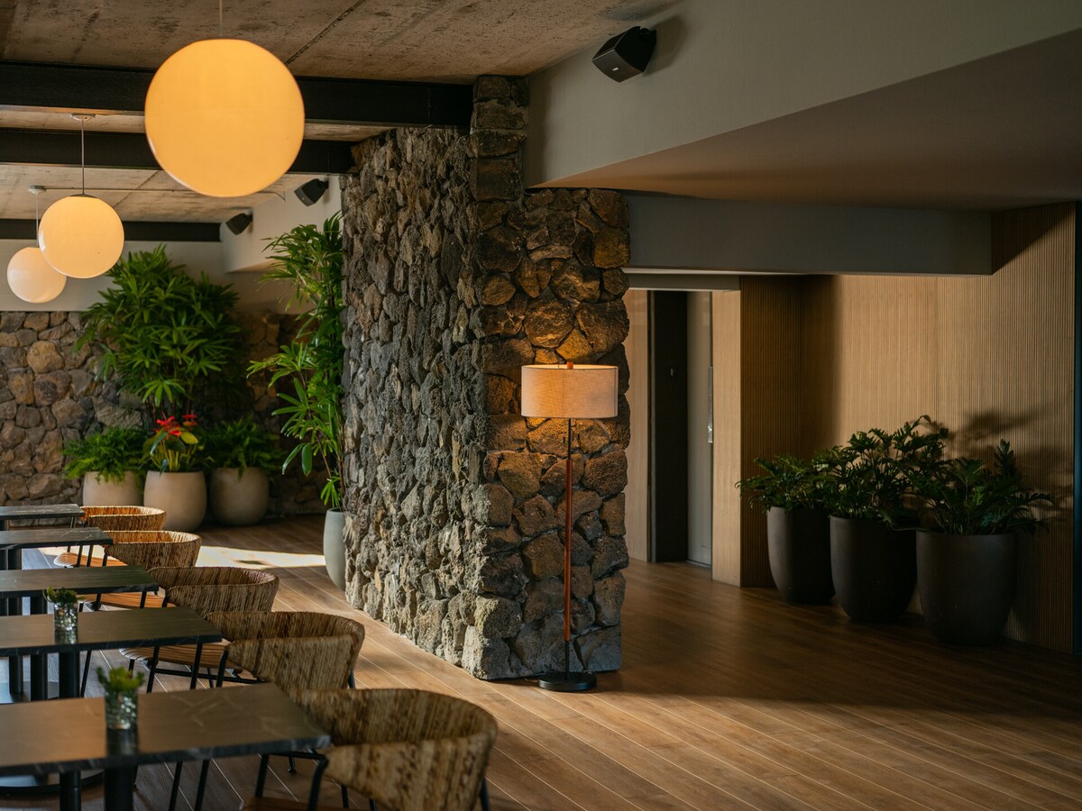 An open dining area features wooden flooring and a curved stone wall adorned with lush greenery in large planters. Soft lighting is provided by circular pendant lamps and a standing lamp, creating a relaxed environment. Tables and chairs are arranged neatly throughout the space.