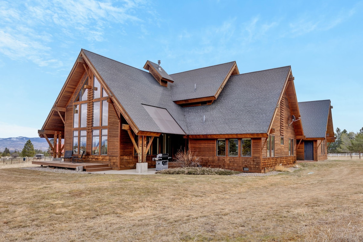 A large wooden home with a spacious deck is showcased under a clear blue sky. Featuring an A-frame design, the structure presents multiple gabled roofs and large windows, allowing natural light to flood the interior. A vast grassy area surrounds the property.