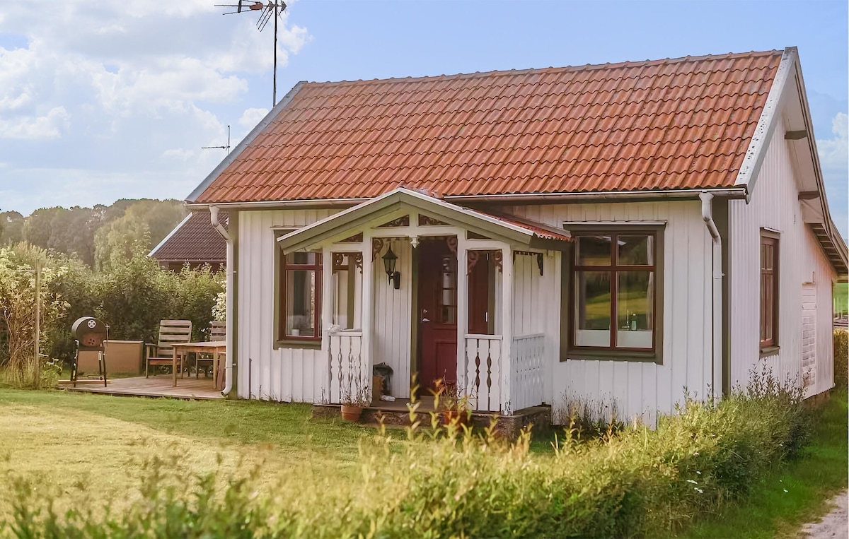 A charming cottage is depicted with a red-tiled roof and white wooden siding. A small porch features a welcoming entrance. Outdoor seating is visible, along with a grill, surrounded by well-maintained grass and greenery.