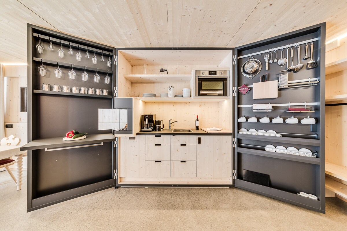 An organized kitchen is presented with cabinetry featuring glassware displayed prominently. The central space includes a compact countertop, an oven, and a coffee maker. Shelves hold various kitchen essentials, while ample light enhances the wood paneling and modern appliances.