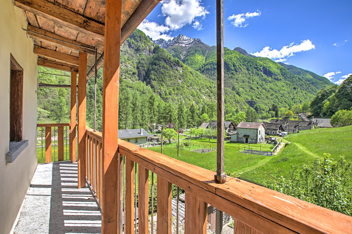 A wooden balcony offers stunning views of the surrounding mountainous landscape and lush greenery. The vibrant blue sky is dotted with fluffy white clouds, while a quaint village with simple structures can be seen in the distance, blending harmoniously with nature.