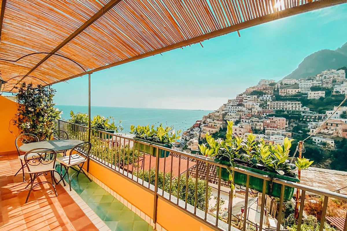 A spacious balcony overlooks the stunning Amalfi coastline, featuring a table and chairs for outdoor dining. Lush greenery borders the railings, complementing the vibrant colors of the landscape. The scene captures the charming houses built into the hillside, with the sea visible in the background.