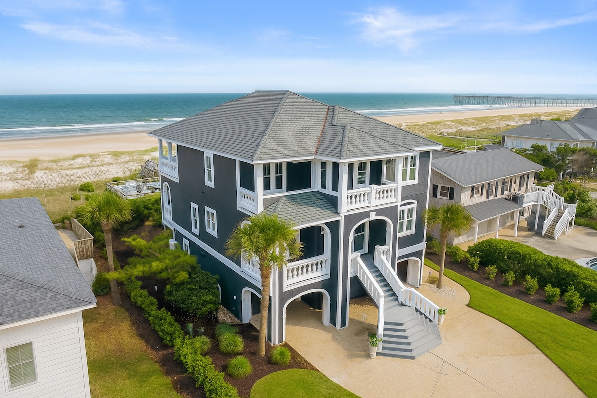 An oceanfront home with a spacious design is showcased against a backdrop of the coastline. Lush landscaping surrounds the property, with palm trees and greenery enhancing the outdoor aesthetic. The house features multiple balconies and stairs leading to the entrance, highlighting its inviting architecture.