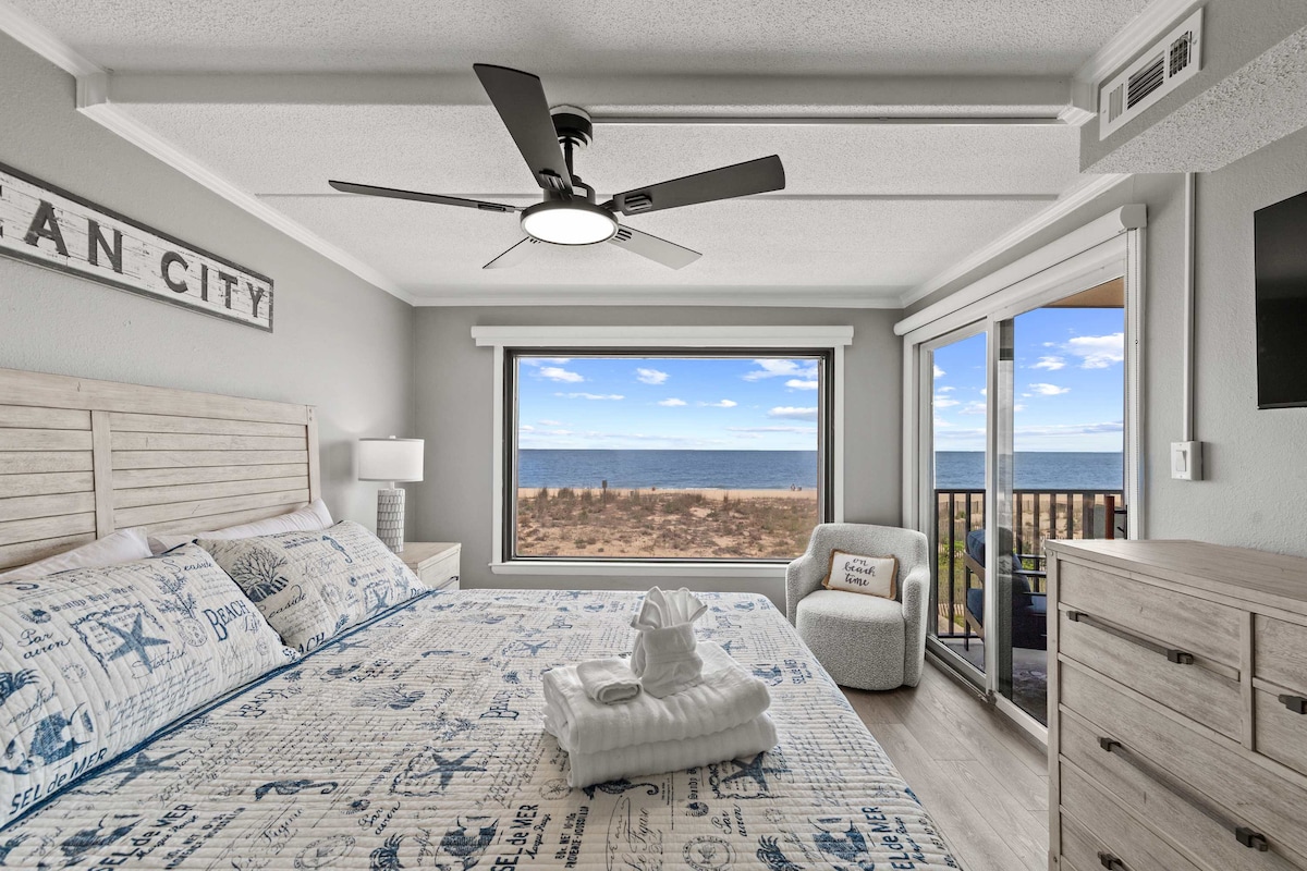 A spacious bedroom features a large window showcasing views of the ocean. A king-sized bed is centered beneath the fan, adorned with a nautical-themed quilt. A comfortable chair sits in the corner, and sliding glass doors lead to a private balcony.