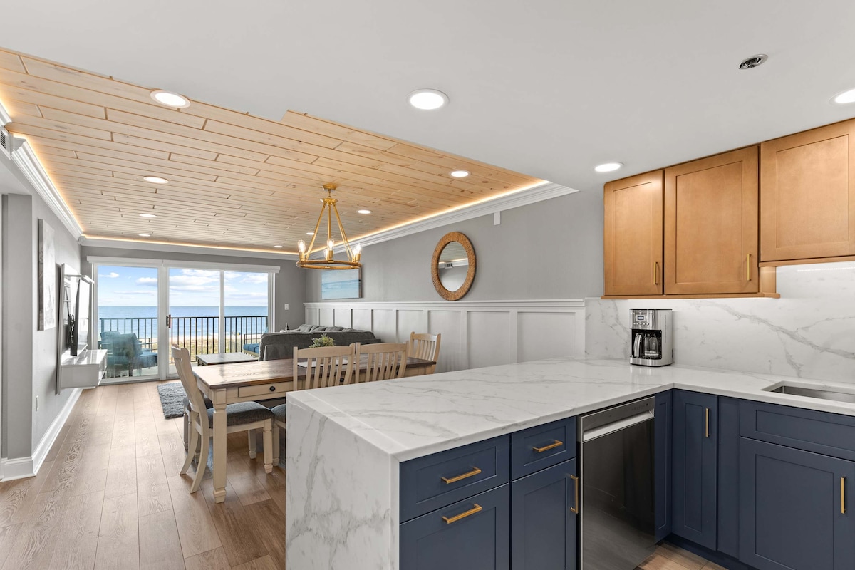 The kitchen features marble-patterned quartz countertops and two-tone cabinetry, blending elegance with functionality. A spacious peninsula provides additional workspace. The adjacent dining area with a wooden table seats six, and large windows allow natural light to illuminate the space while offering views of the ocean.