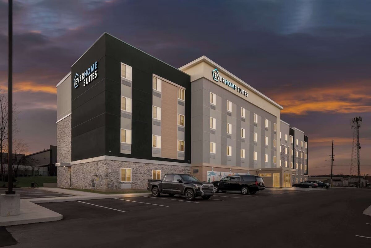 A modern extended-stay property is shown, featuring a five-story building with a mix of green and light-colored exteriors. The softly illuminated entrance is highlighted as dusk approaches, along with ample parking spaces in front of the structure.