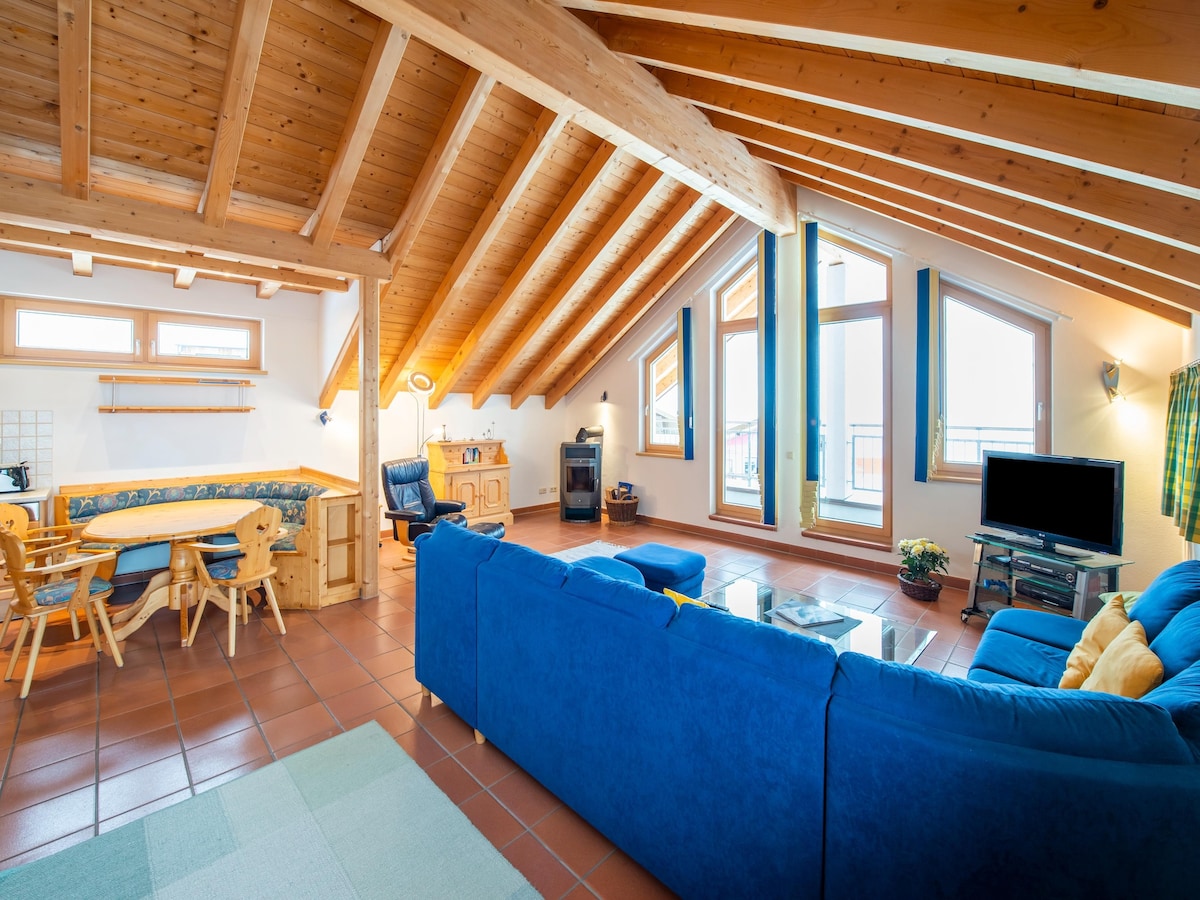 The spacious living room features a blue sectional sofa facing a television. Large windows allow for natural light and offer views of the outdoors. A dining area with a circular table and wooden chairs is also present. The room is complemented by sloped wooden ceilings.