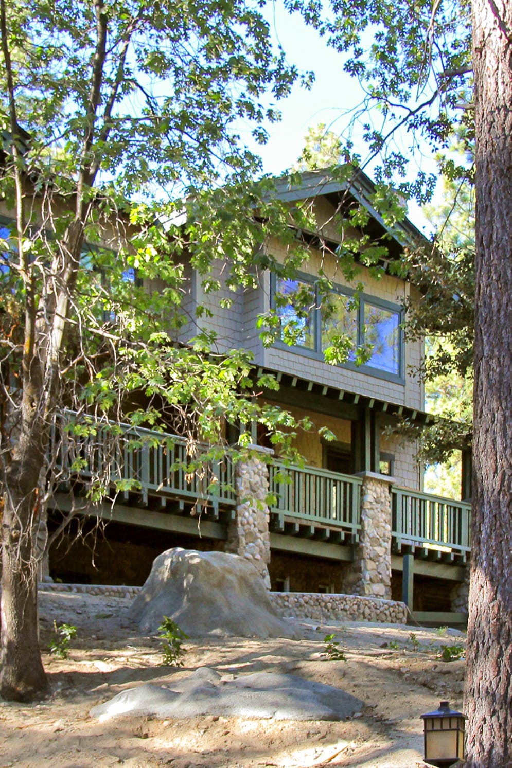 A multi-level lodge is surrounded by trees, showcasing a stone and wood exterior. The expansive front porch is supported by wooden railings. Large windows offer glimpses of the interior, while natural elements blend seamlessly with the structure.