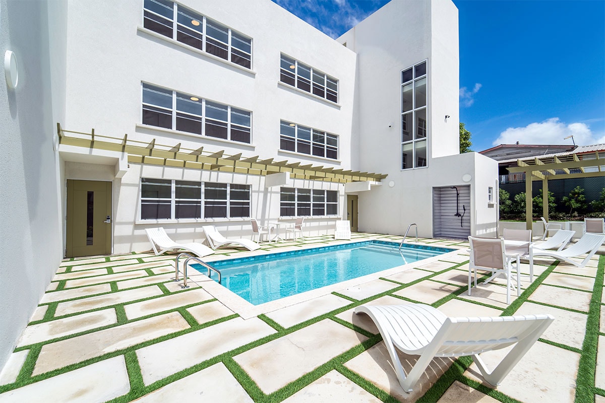 A shared swimming pool is framed by modern white buildings, with lounge chairs positioned around the pool deck. The area is designed with paved tiles and artificial grass, creating a clean and inviting outdoor space. Large windows allow natural light to fill the surroundings.