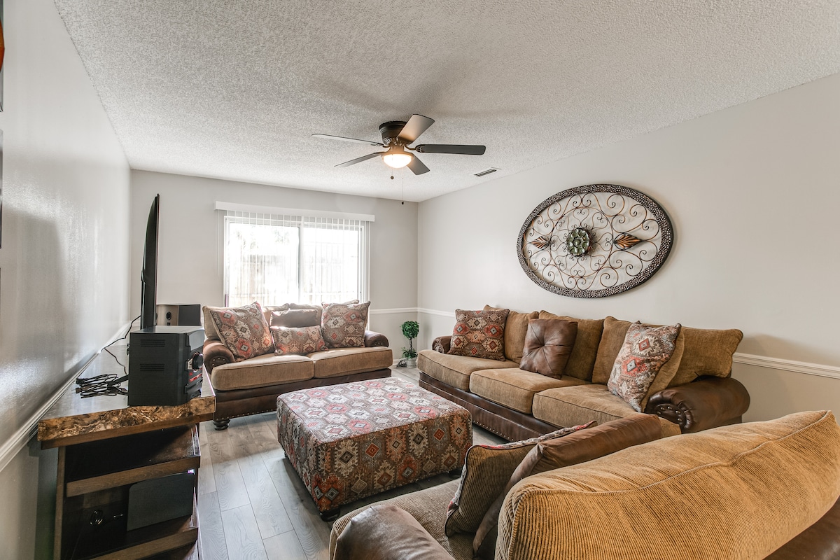 The living area features a mix of brown leather and beige fabric sofas, arranged to promote conversation. A patterned ottoman is positioned at the center. Natural light enters through a window covered by sheer curtains, and an intricate wall decor piece adds visual interest.