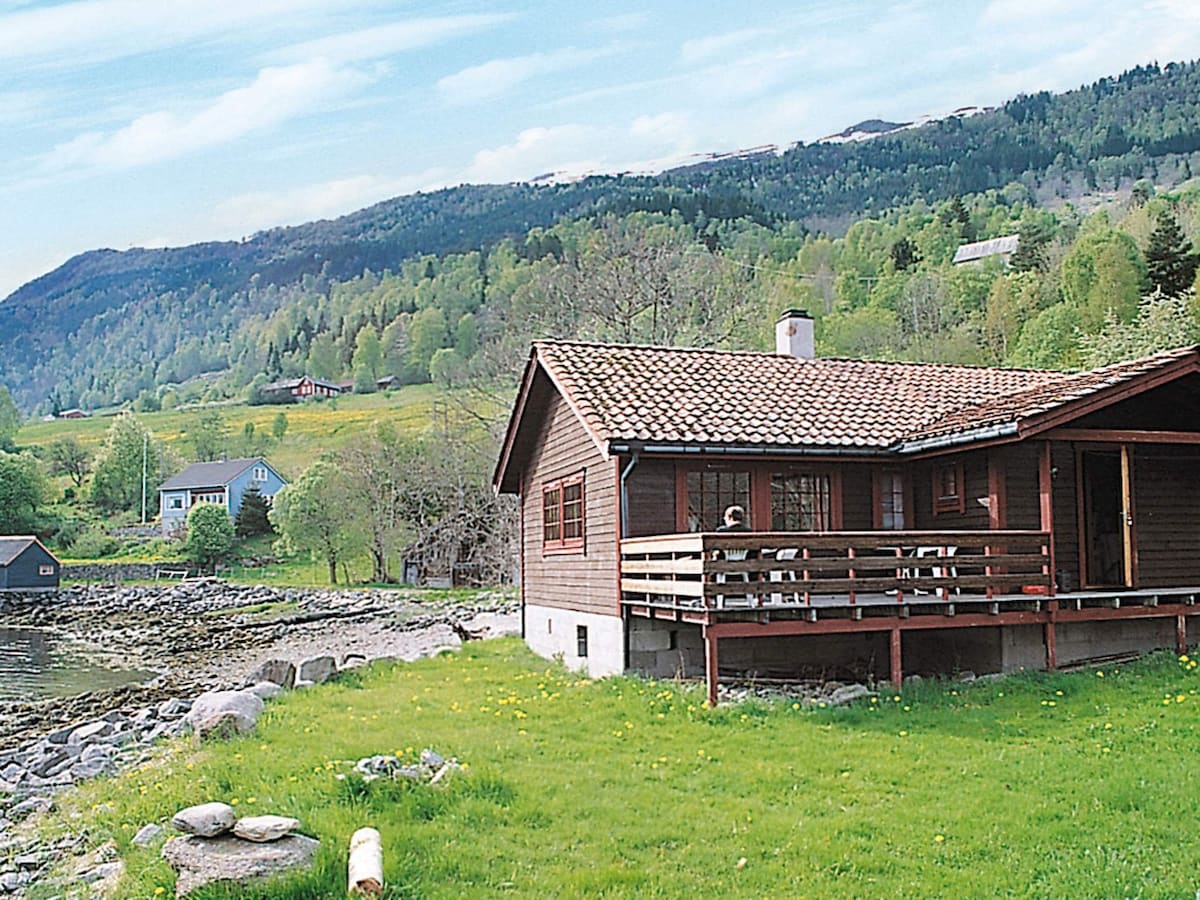 A charming holiday cottage features a wooden exterior and a tiled roof, situated near the fjord. A spacious terrace wraps around the front, overlooking a grassy area and rocky shoreline. Lush green hills dotted with trees rise in the background, creating a serene landscape.